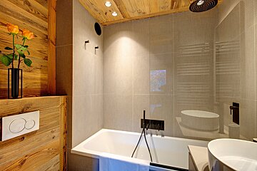 A modern bathroom with a wooden chalet-style ceiling and wall, light tiled shower area, white bathtub, and contemporary black fixtures. Orange roses add color.