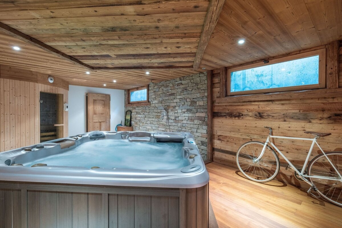 A rustic spa room with a bubbling hot tub, sauna, stone wall, and wooden interior. A white bicycle leans against a wood-paneled wall, illuminated by blue-tinted windows.