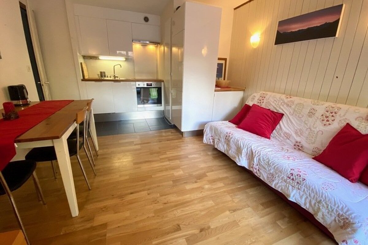 Cozy studio apartment with light wood floors, a dining table with a red runner, a white kitchen, and a sofa with red pillows.