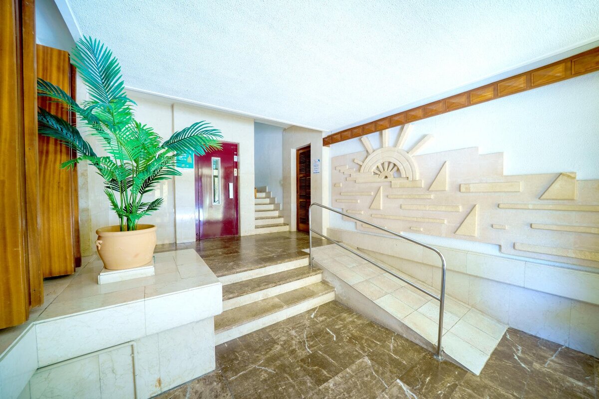 A bright building lobby with a large potted palm, marble floors, stairs, a red elevator door, and geometric sun wall art on the right.