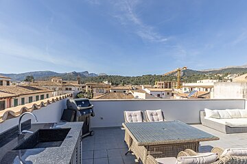 A sunny rooftop terrace with an outdoor kitchen, grill, dining table, and sofa, overlooking a town with terracotta roofs and distant mountains.