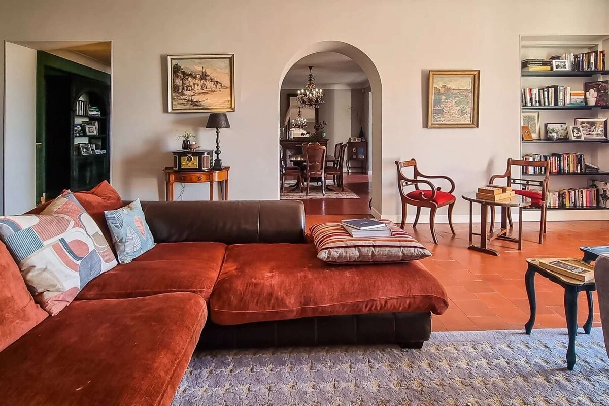 A cozy, traditional living room with a large L-shaped sofa, terracotta floor, and an arched doorway leading to a dining room. Features include classic decor and bookshelves.