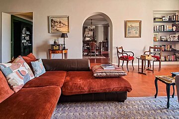 A cozy, traditional living room with a large L-shaped sofa, terracotta floor, and an arched doorway leading to a dining room. Features include classic decor and bookshelves.