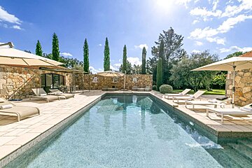 A serene outdoor swimming pool with sun loungers, stone walls, and tall cypress trees under a bright, sunny sky.