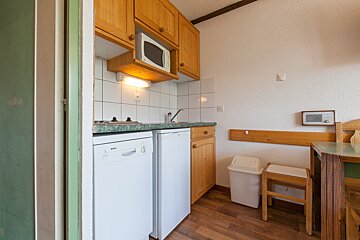 A compact kitchen with white cabinets, dark countertops, built-in oven, sink, and a window looking into another room. Dark wall and wooden floor.