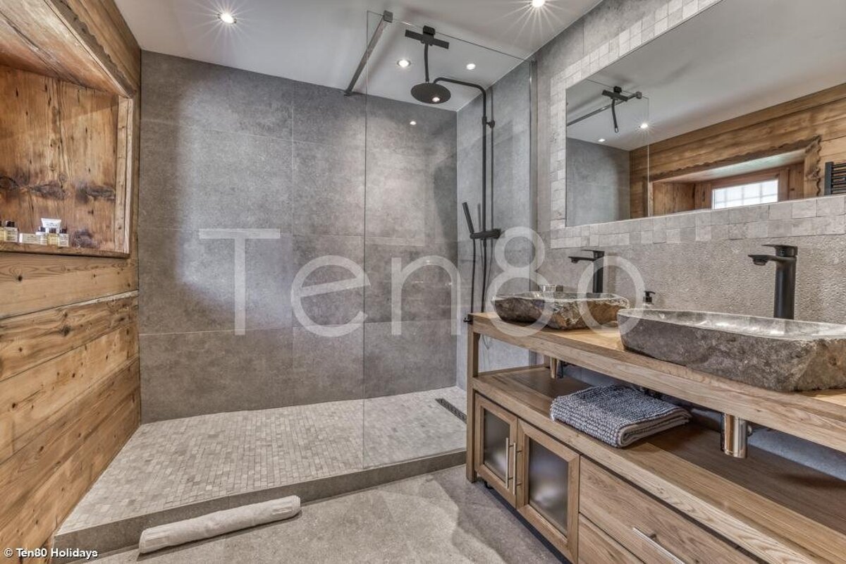 Modern bathroom with a glass walk-in shower, grey tiles, wooden wall panels, and a vanity with two natural stone sinks.