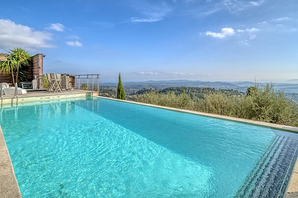 An outdoor infinity pool with turquoise water overlooking a scenic valley of rolling green hills under a blue sky.