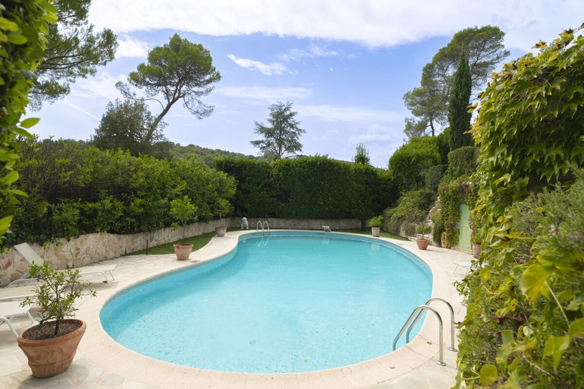A curved swimming pool surrounded by lush green hedges, trees, potted plants, and lounge chairs in a private backyard.