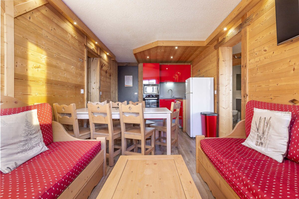 Cozy chalet apartment with warm wood paneling, a vibrant red kitchen, dining table, and two red daybeds, creating an inviting ski lodge feel.