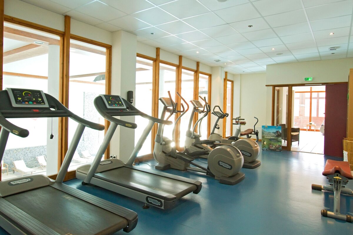 A bright, modern gym with treadmills, ellipticals, and exercise bikes, featuring large windows overlooking an outdoor area and a blue floor.