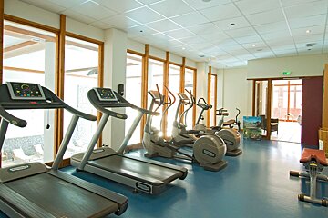 A bright, modern gym with treadmills, ellipticals, and exercise bikes, featuring large windows overlooking an outdoor area and a blue floor.