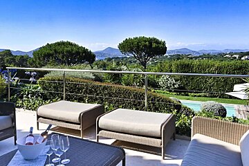 Sunny patio with wicker seating and wine, offering a panoramic view of a coastal landscape, green hills, lush trees, and a distant pool.