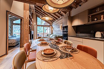 Modern chalet dining area with a set wooden table, unique pendant lights, a sleek kitchen, and a glimpse of a bedroom to the left.