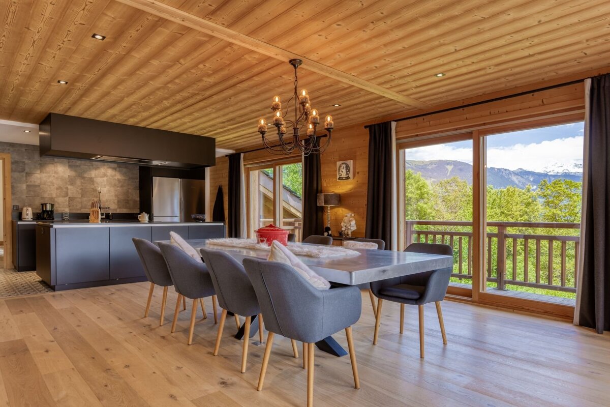Chalet-style open-plan kitchen and dining room with wooden interiors, modern grey kitchen, dining table, and a scenic mountain view through large windows.