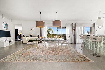 Bright, modern dining area with a glass table on a large rug, pendant lamps, bar stools, and sliding doors to a scenic view.