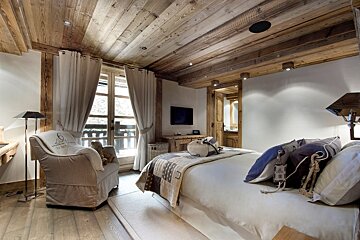A cozy chalet bedroom with a rustic wooden ceiling, large bed, armchair by a balcony door, and a warm, inviting ambiance.