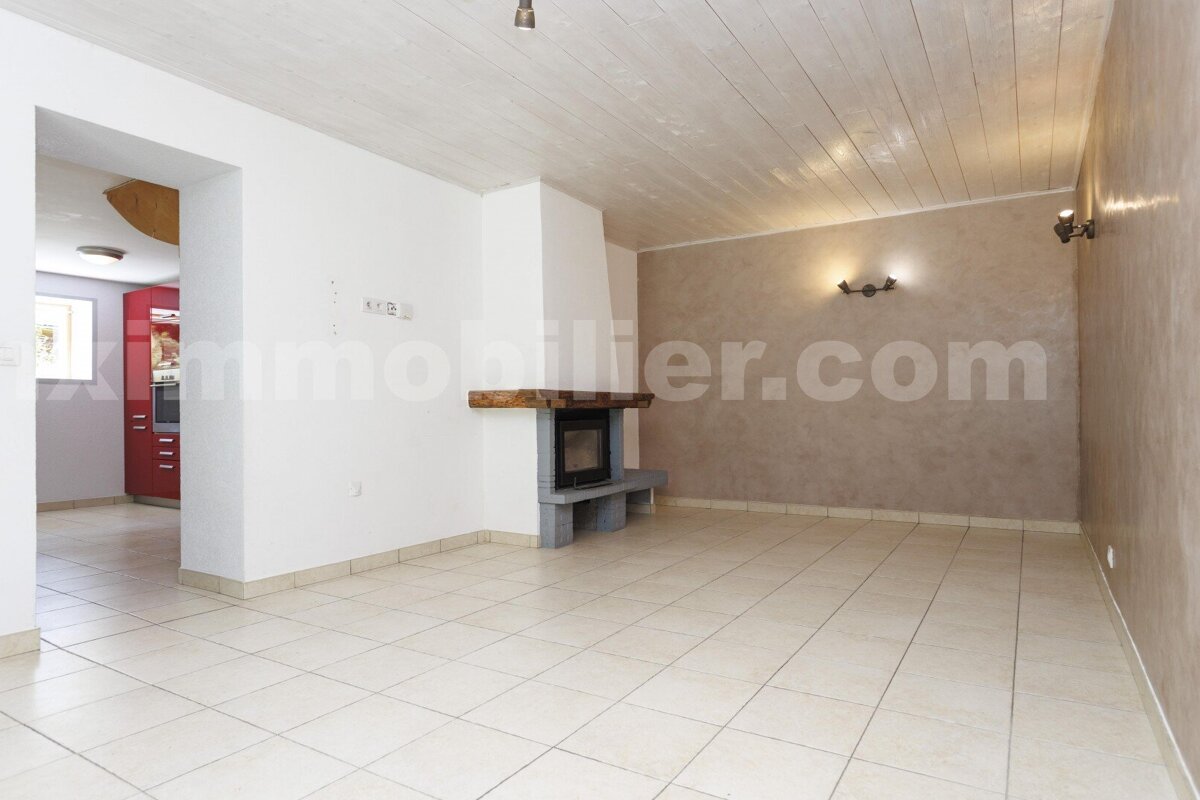 Empty room with light tiled floor, white and beige walls, a fireplace, and a wooden plank ceiling. A red kitchen is visible through an archway.
