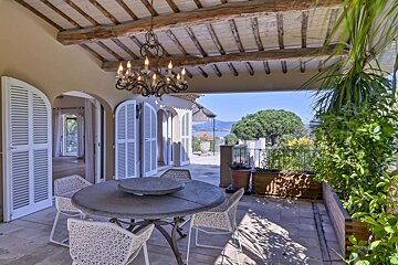 A chic covered patio with a dining set, white shutters, and a chandelier, overlooking a coastal landscape with a distant sea and lush trees.