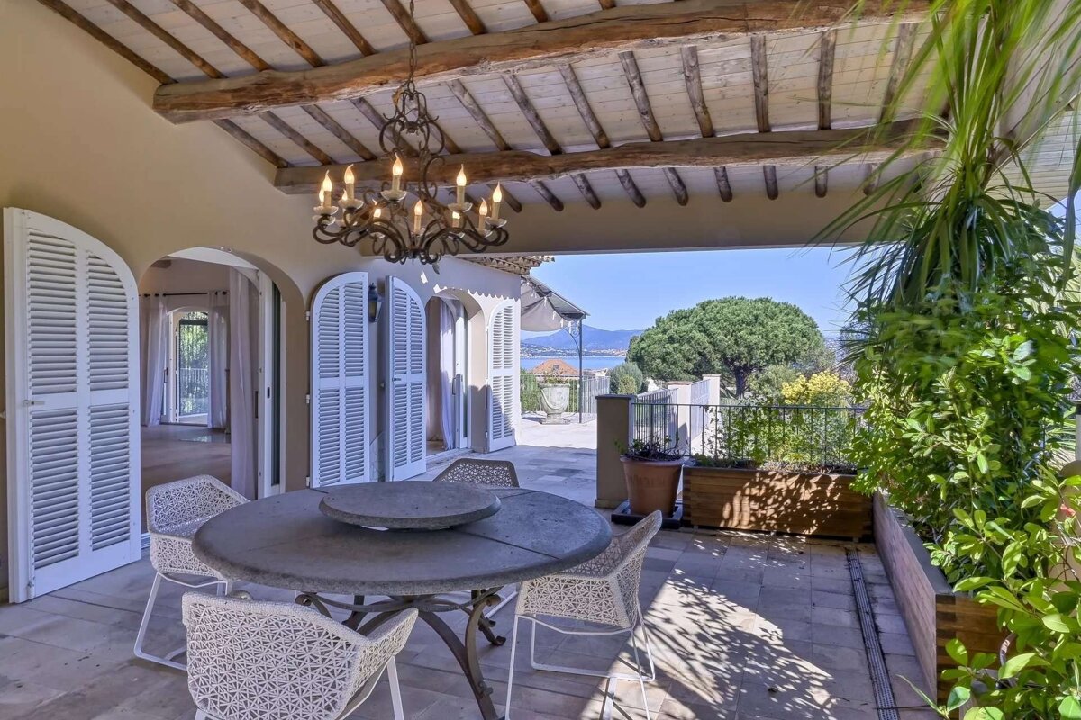 A charming, covered Mediterranean-style terrace with a dining set, chandelier, and lush plants, overlooking a beautiful sea and distant hills.