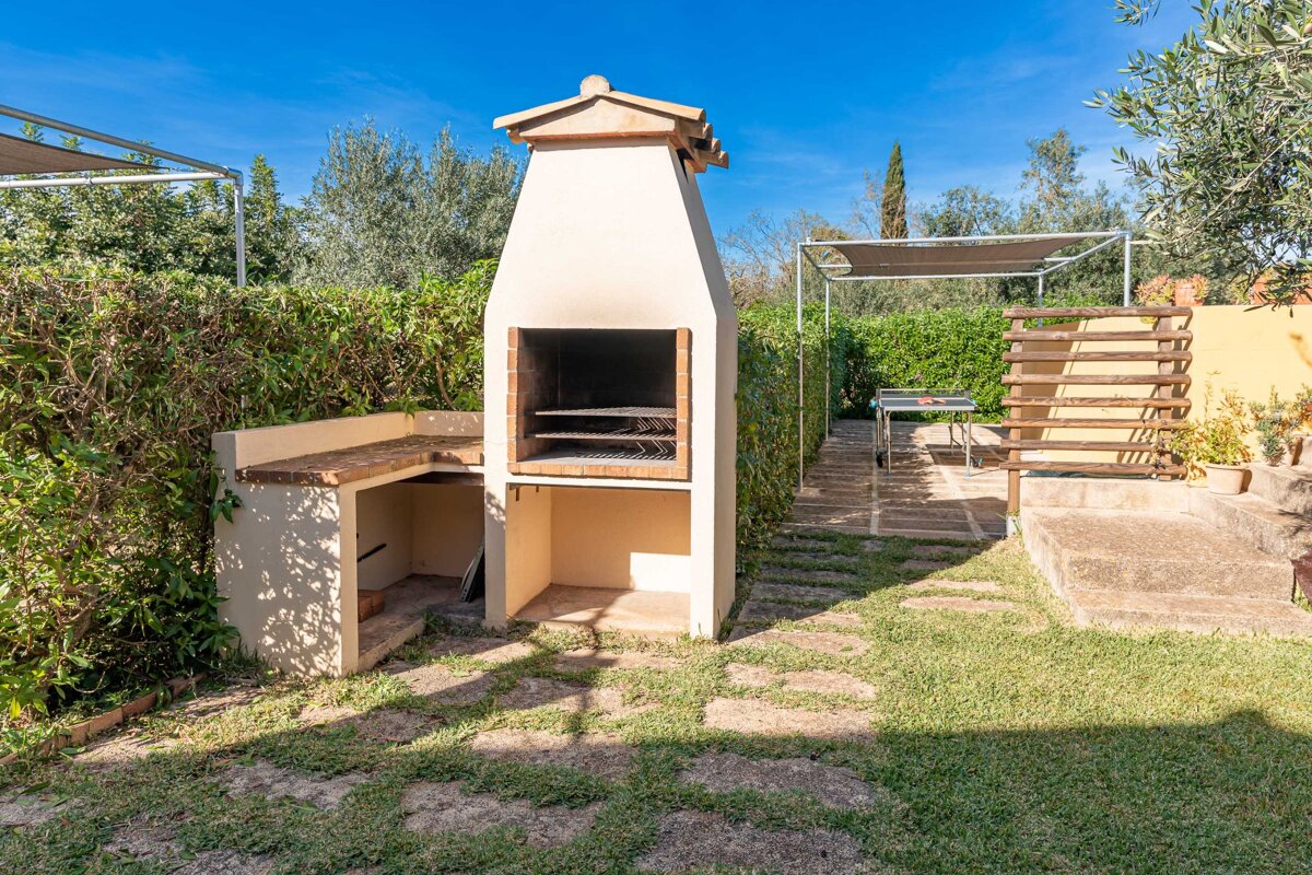 An outdoor built-in barbecue area with a stone path, green grass, lush hedges, and a clear blue sky, set up for al fresco dining.