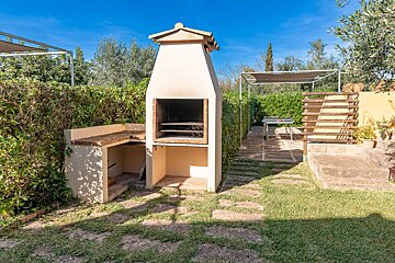 An outdoor built-in barbecue area with a stone path, green grass, lush hedges, and a clear blue sky, set up for al fresco dining.