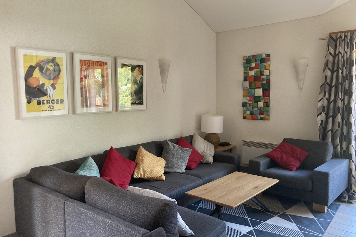 A cozy modern living room with a gray L-shaped sofa, vibrant cushions, and an armchair. Features art posters, a mosaic wall hanging, and a geometric rug.