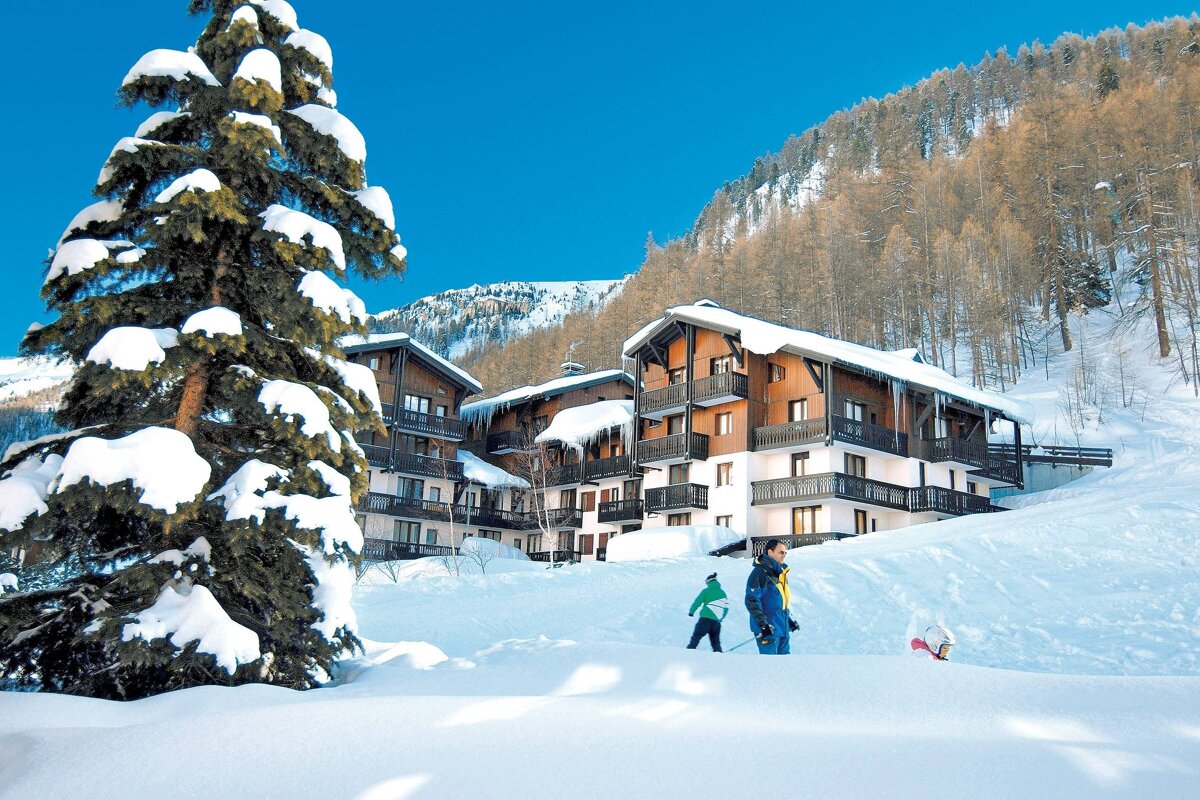 A snowy alpine village features charming chalets, a large snow-laden pine, and people playing in deep snow. Forested mountains meet a clear blue sky.