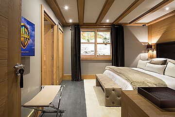 A cozy, modern bedroom with wooden ceiling beams, a large bed, window, TV, and neutral decor. Keys hang by the door.