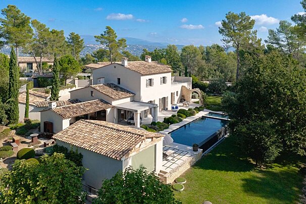 Aerial view of a sprawling white Mediterranean-style villa with a dark pool, lush gardens, and abundant trees under a bright blue sky.