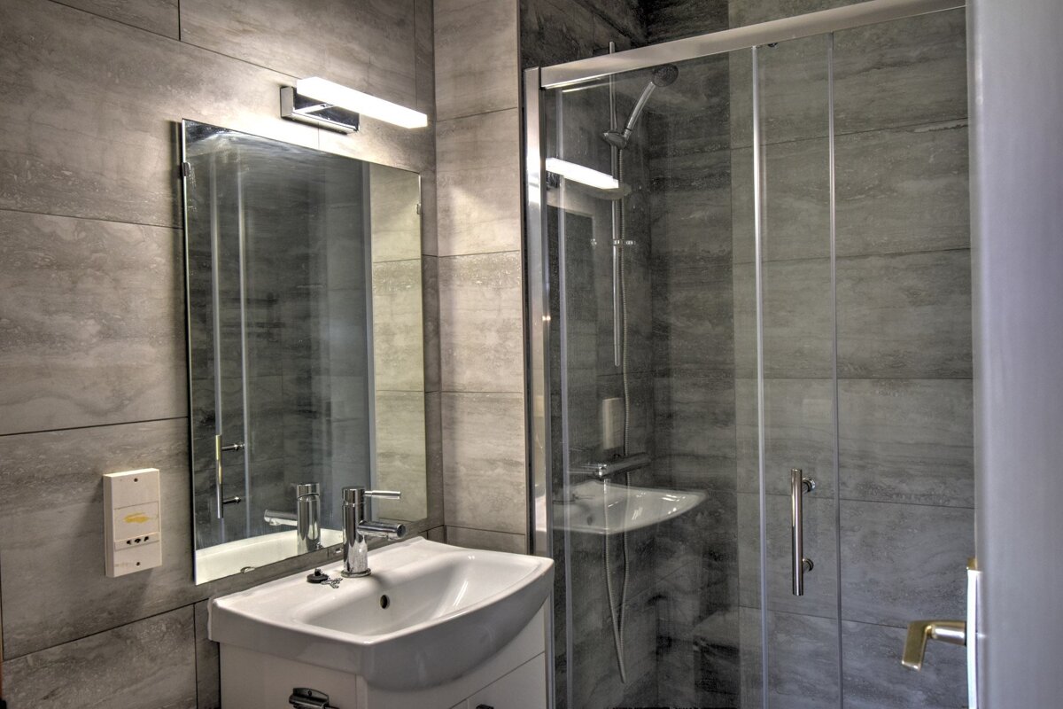 A modern bathroom with gray tiled walls, a white sink, an illuminated mirror, and a glass shower enclosure.