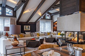 Luxurious chalet living room with vaulted wood-beamed ceiling, plush sofa, modern fireplace, geometric tables, and an adjacent dining area.