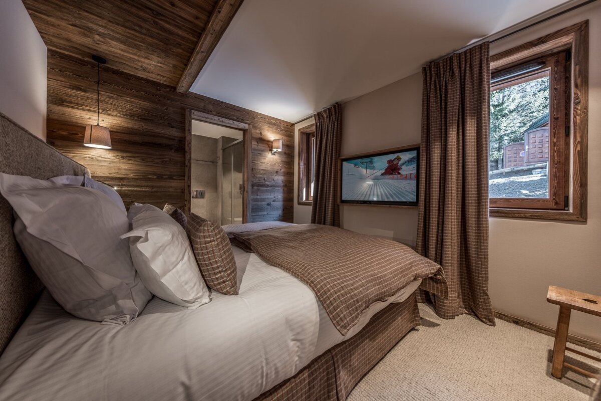 Cozy chalet bedroom with rustic wooden walls and ceiling, a large bed, TV, window, and attached bathroom. Brown and white decor.