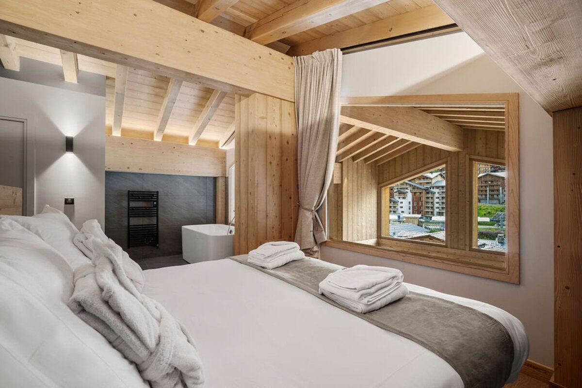 Modern chalet bedroom with wooden beams, a large white bed, an open bathtub area, and a window overlooking an alpine village.