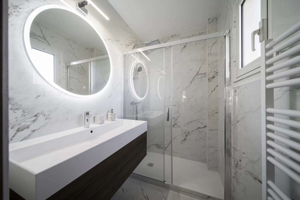 A sleek modern bathroom featuring white marble walls, a backlit oval mirror above a floating vanity, and a clear glass shower enclosure.