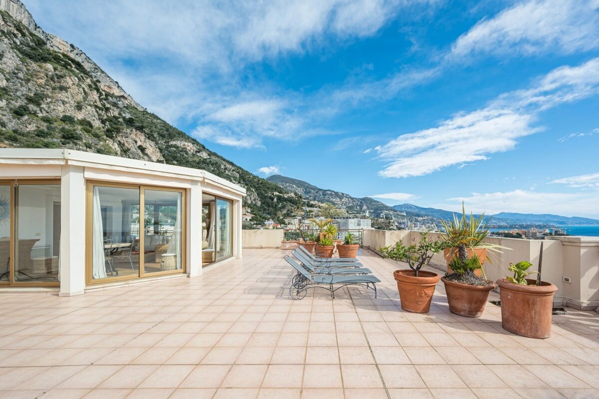 Rooftop terrace with panoramic views of mountains, city, and sea. Features a glass-enclosed room, lounge chairs, and potted plants under a blue sky.