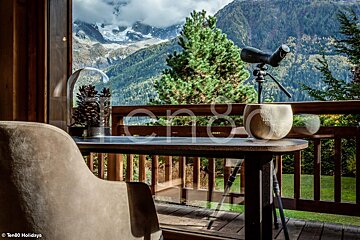 A cozy balcony offers stunning mountain views with snowy peaks, lush forests, a spotting scope, and pinecone decor on a wooden table.