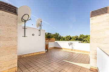 A spacious rooftop terrace with beige tiling, white walls, satellite dishes, and a view of green trees under a clear blue sky.