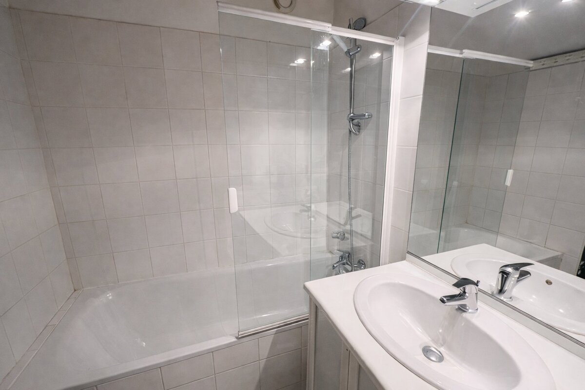 A bright, tiled bathroom with a bathtub, glass shower screen, and a white sink with a mirror.