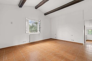 Empty room with white walls, terracotta tiled floor, exposed wooden beams, and a window with security bars above a white radiator.