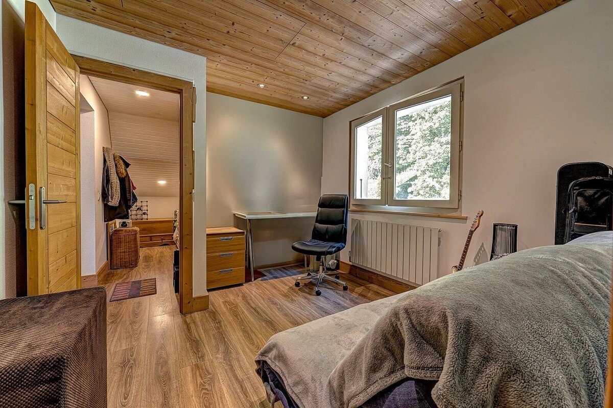 Cozy room with wood ceiling and floor, featuring a bed, desk, office chair, window with outdoor view, and a guitar, with an open doorway.