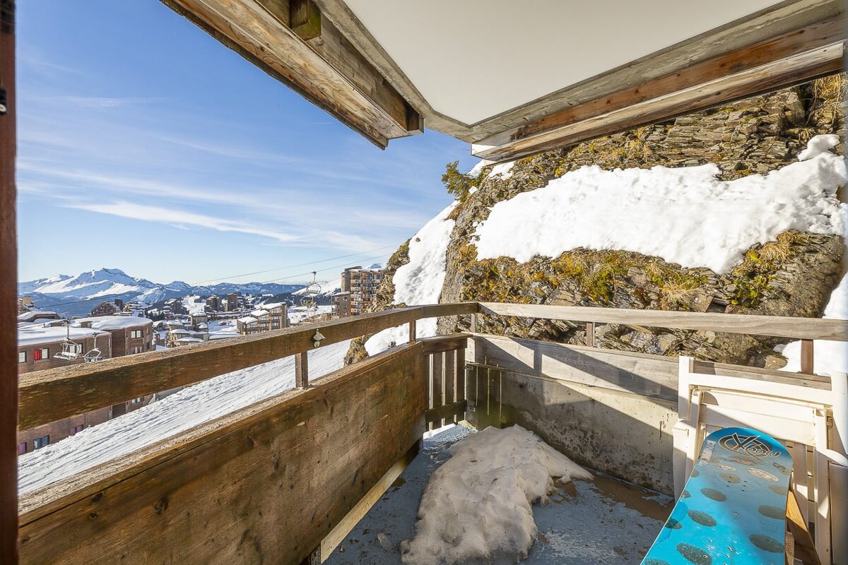 A snowy balcony with a snowboard overlooks a mountain ski resort and village, framed by a rocky, snow-covered cliff under a clear blue sky.