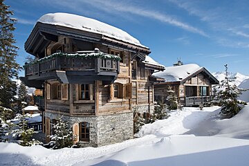 A snow-covered wooden chalet with a balcony, surrounded by pine trees and snow, with distant mountains under a clear blue sky.
