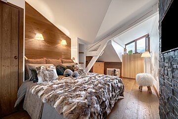 Cozy chalet bedroom with faux fur bed, wood paneling, angled ceiling, and mountain views through a window, exuding a warm lodge feel.