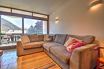 A cozy living room with a grey L-shaped sofa, striped rug, and a large window offering a scenic view of a mountain village and peaks.