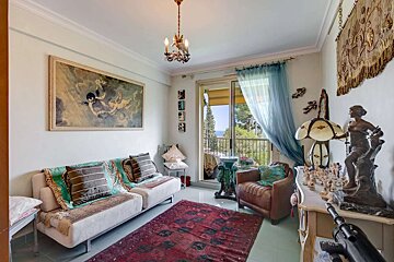 An eclectic living room with a red rug, ornate decor, a sofa, and an armchair, leading to a balcony with a distant sea view.