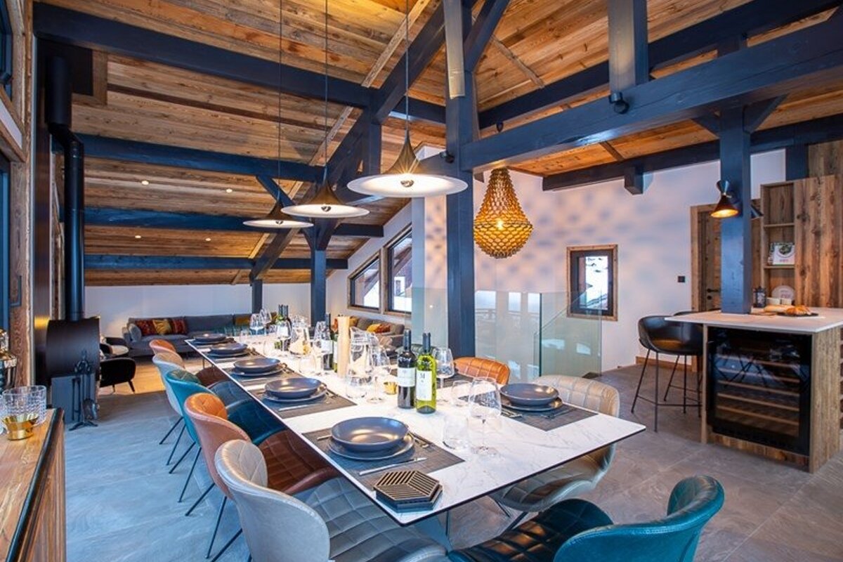 A modern chalet dining room with a long set table, colorful chairs, and exposed wooden ceiling beams.