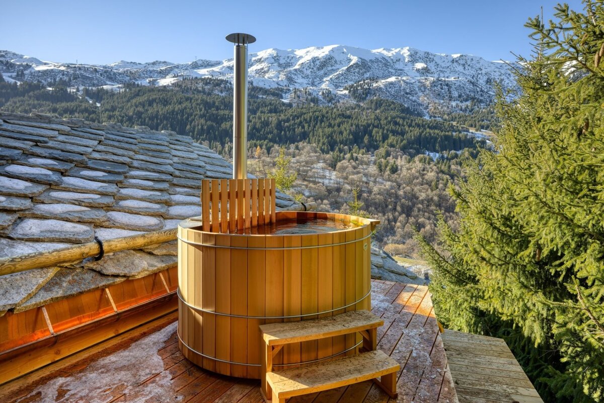 A rustic wooden hot tub overlooks a stunning panorama of snow-capped mountains and a pine forest under a clear sky.