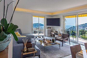 A bright living room with a blue sofa, TV, and chess set. Sliding doors open to a balcony with yellow awnings and stunning sea & mountain views.