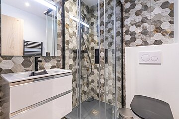 A modern bathroom featuring hexagonal grey and brown tiled walls, a white floating vanity with a black faucet, a glass shower, and a toilet.