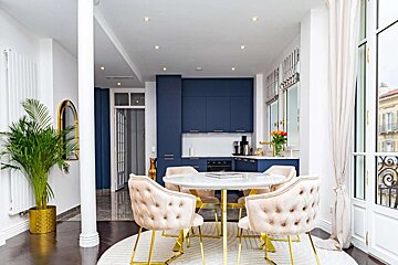 A stylish open-concept kitchen and dining area with dark blue cabinets, a marble table, and tufted pink chairs with gold accents. Modern and elegant.
